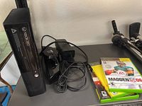 Xbox 360 console standing vertically with power cables and three games stacked next to it on a shelf.
