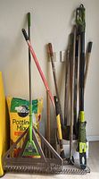 Full view of the collection including rakes, shovels, axe, and electric pole chainsaw alongside potting mix bags.