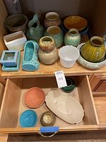 Various handmade pottery vases in green, beige, turquoise, caramel tones, and a white hobnail planter, displayed on a drawer and cabinet shelf