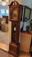 Full view of tall grandfather clock showing wooden case, bonnet top with finials, and glass paneled pendulum door.