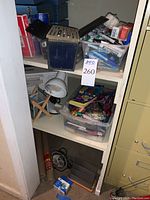 Shelf with bins containing various pens, markers, glue bottles, tape, and other office supplies. A small white gooseneck lamp is positioned on the shelf along with a black desk fan and other miscellaneous items.