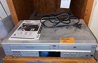 Front of Panasonic VHS/DVD player unit with power cord and manual on top. Includes VHS slot and DVD slot with control buttons and input jacks visible.