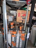 Thirteen threaded steel bars with orange clamps standing vertically on a rack, showing clamp mechanisms and bars.