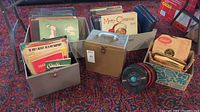 Boxes containing vintage vinyl records and music books on carpeted floor, records include various genres and many in original sleeves.