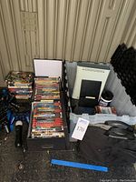 Photo of PlayStation 2 console, Xbox 360 console, wired controllers, and games/DVD cases stored on the ground and in bins.