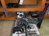 Xbox 360 console, wired and wireless controllers, and stacks of PlayStation 2 and Xbox 360 game cases on shelf