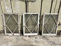 Front view of three diamond pane windows with white frames placed upright against a container, visible chipped paint on frames, two windows have cracked glass bottoms.