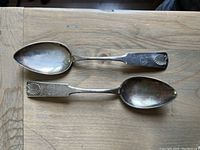 Two Finnish silver serving spoons laid side by side on a wooden surface, showing bowl and handle details including shell motif and engraving.