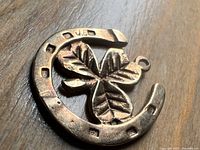 Close-up photo of the gold charm, showing the horseshoe and clover design with hallmark stamps visible on the horseshoe's edge.