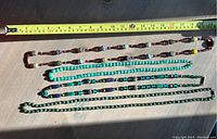 Four necklaces laid out next to measuring tape showing lengths between 14 and 19 inches, beads in various shapes and colors including black and red wooden beads, turquoise beads, blue-green glass beads, and green stone beads.