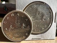 Close-up photo of the reverse sides of the 1950 and 1951 Canadian silver dollar coins showing the canoe design with two figures paddling and maple leaves.