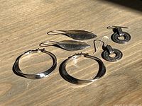 Photo showing three pairs of assorted silver earrings including leaf-shaped drops, large wavy hoops, and small circular dangle earrings.