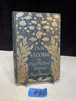 Front cover showing dark hardcover with detailed gold embossed floral design and title Our Village by Miss Mitford.