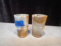 Two polished onyx glasses standing side by side on a light surface with a dark background. Each glass shows natural stone color variations from cream to brown with a smooth sheen.