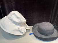 Two vintage ladies hats displayed side-by-side on a flat surface - a white textured fabric hat with a bow and a black woven hat with a structured brim.