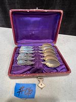Set of six antique sterling silver spoons arranged inside their original red case lined with purple satin, showing detailed ornate handles and smooth spoon bowls.
