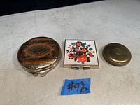 Three closed compact cases: a copper-toned decorative metal compact, a square floral decorated compact, and a small round gold-toned compact.