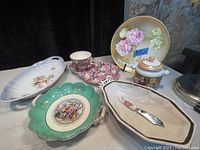 Full lot of seven various vintage ceramic and porcelain pieces with floral and classical designs laid out on a table.