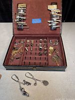 Brown faux leather display case opened to reveal multiple compartments holding vintage tie clips, tie pins, cufflinks, and money clips. Two metal key chains with clips are placed outside the case on a light surface.