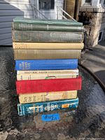 Photo of a stack of 11 antique and vintage hardcover books showing spines and top edges on outdoor table with normal age wear.