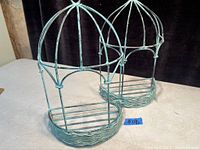 Pair of metal plant hangers with open cage design and woven bases. Both have a rounded top and patina finish.