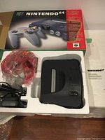 Nintendo 64 console in styrofoam tray with red translucent controller in plastic, AC adapter, and original box.