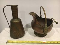 Two vintage solid brass wares including a tall etched floral brass jug and a brass coal scuttle with ceramic handle, shown with measuring tape for size reference.