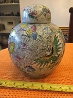 Front view of vintage Oriental jar with lid showing colorful butterfly and floral patterns along with measuring tape at base.