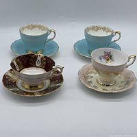 Four assorted fine china tea cups with matching saucers, including a blue set, a dark burgundy and gold decorated set, and two pink floral sets, arranged on white background