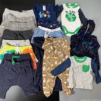 Full lot of assorted infant clothing items including pants, bodysuits, tops, and pajamas shown laid out on a black surface.