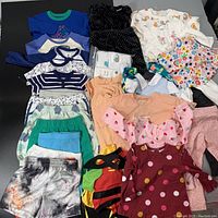 Overview of assorted infant clothing including tops, dresses, pants, and crib sheet laid out