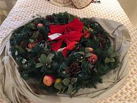 Top view of a 30-inch diameter artificial Christmas wreath on a bed, decorated with pinecones, red faux apples, green leaves, red berries, and three red velvet bows.