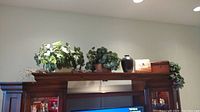 Wide shot of faux greenery plants, decorative boxes, and a dark vase arranged on top of a wooden mantel