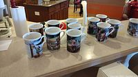 12 Hummel collectible coffee cups arranged on countertop, each with unique artwork representing different months.