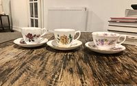 Photo displaying three bone china tea cups and matching saucers placed in a row on a wooden surface, showing their different floral designs.