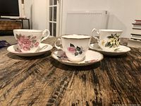 Three tea cups with matching saucers on rustic wooden table, each with floral pattern and gold trim.