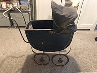 Side view of the black wooden doll carriage with metal frame and fabric hood, showing the curved handle and overall structure.