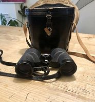 Vintage binoculars with textured grip beside their black leather carrying case with strap, showing overall condition and accessories.