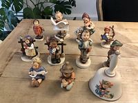 Photo showing 10 Hummel figurines arranged on a table, featuring children in various activities and poses. Some have visible small chips.