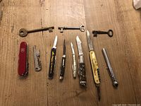 Top view showing all items arranged on wood surface including 5 pen knives, Swiss Army knife, bottle opener, tire gauge, and 3 skeleton keys