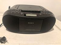 Front view of a black Sony boombox showing dual speakers, cassette player door with Sony branding, control buttons, and LCD display.