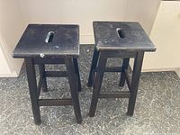 Pair of black wooden bar stools side by side on floor, visible scratches and wear on seats.