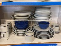 Wide shot showing stacked white plates with blue dot pattern, blue ceramic bowls, white mugs with blue rim, and a white metal shelving rack.