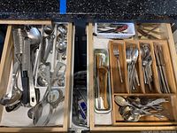 Top view of two open drawers showing assorted kitchen utensils and flatware in a wooden organizer and loose in drawer, including graters, ladles, tongs, forks, spoons, and knives.