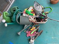Photo showing multiple green and gray plastic watering cans, a galvanized metal watering can, garden hose, gloves, and garden hand tools piled inside a plastic tub on a blue floor.