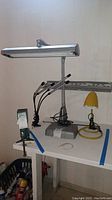 Photo of three lights on a white table, showing the silver 4-head clamp light, silver desk lamp, and yellow desk lamp.