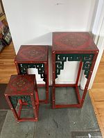 Three Asian-themed nesting tables lined up by size on stone floor, showing red lacquer finish and green fretwork legs