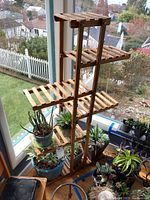 Wooden multi-tiered plant stand with plants on slatted shelves near window.