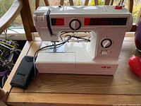 Front and left side view of Elna 1010 sewing machine showing stitch selection dial, secondary dial, stitch pattern guide, and foot pedal on wooden surface.