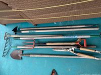 Top-down view of six yard tools laid out horizontally on blue surface showing full length including handles and working ends.